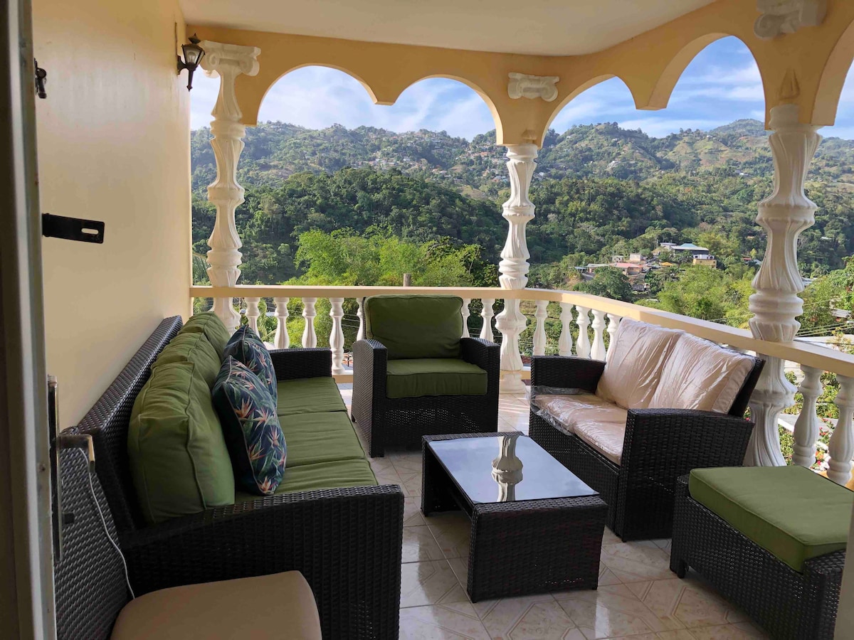 A covered porch features a comfortable seating arrangement with a mix of wicker chairs and a sofa, adorned with colorful cushions. A glass-top coffee table is centered among the seating. Arched columns frame the outdoor space, offering picturesque views of the surrounding hills.