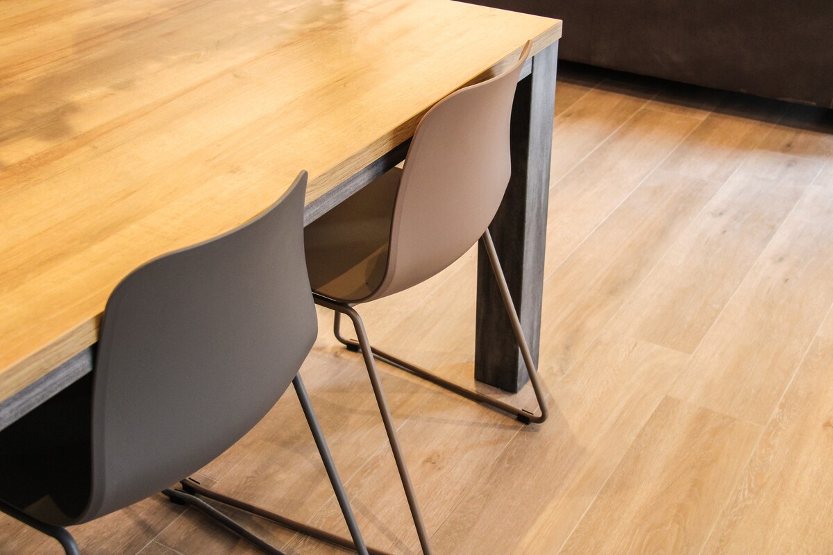 A modern dining table made of wood is featured, supported by sturdy metal legs. Two minimalist chairs in neutral tones are positioned neatly beside the table. A smooth wooden floor complements the furniture, adding warmth to the space.