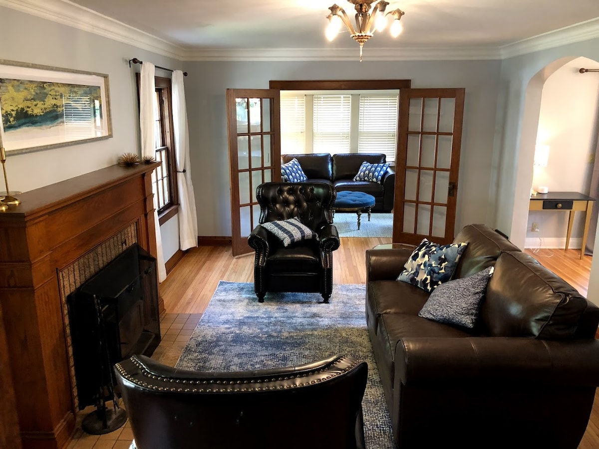 A welcoming living area features a comfortable leather sofa and an armchair, arranged on a soft area rug. A fireplace adds a classic touch, while the large windows allow natural light to illuminate the space. French doors lead to a cozy seating area in the background.