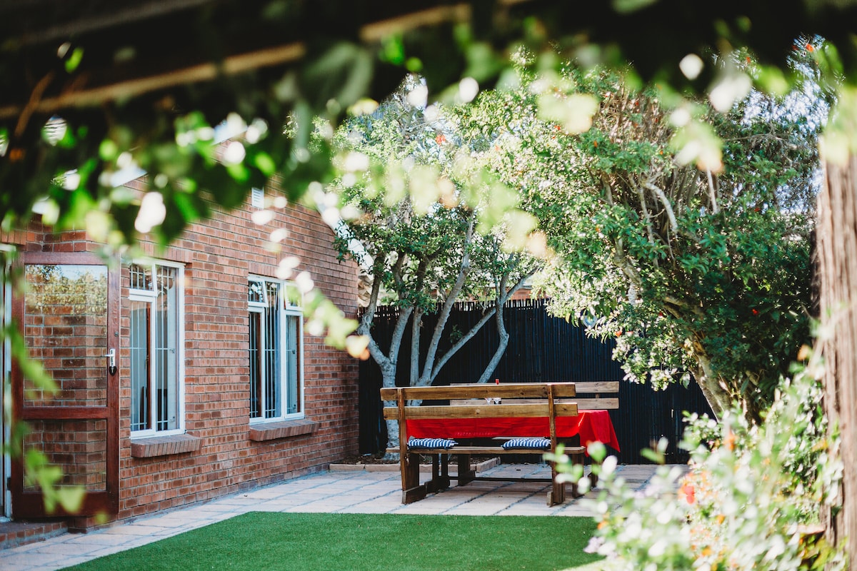 A spacious outdoor dining area is framed by greenery, featuring a wooden table with benches. The brick façade of the cottage is visible in the background, complemented by lush trees and a manicured lawn.