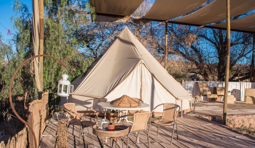 A cozy tent is situated on a wooden deck, surrounded by natural landscapes. A dining table and several wicker chairs are arranged nearby, providing a relaxing outdoor space. Sunlight casts a warm glow, enhancing the inviting atmosphere of this unique accommodation.