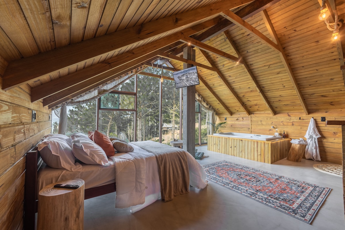 The spacious sleeping area features a wooden ceiling with exposed beams, creating a rustic ambiance. Large windows allow natural light to fill the space, offering tranquil views of the outdoors. A cozy bed is paired with soft linens, and a stylish rug adds warmth to the floor.