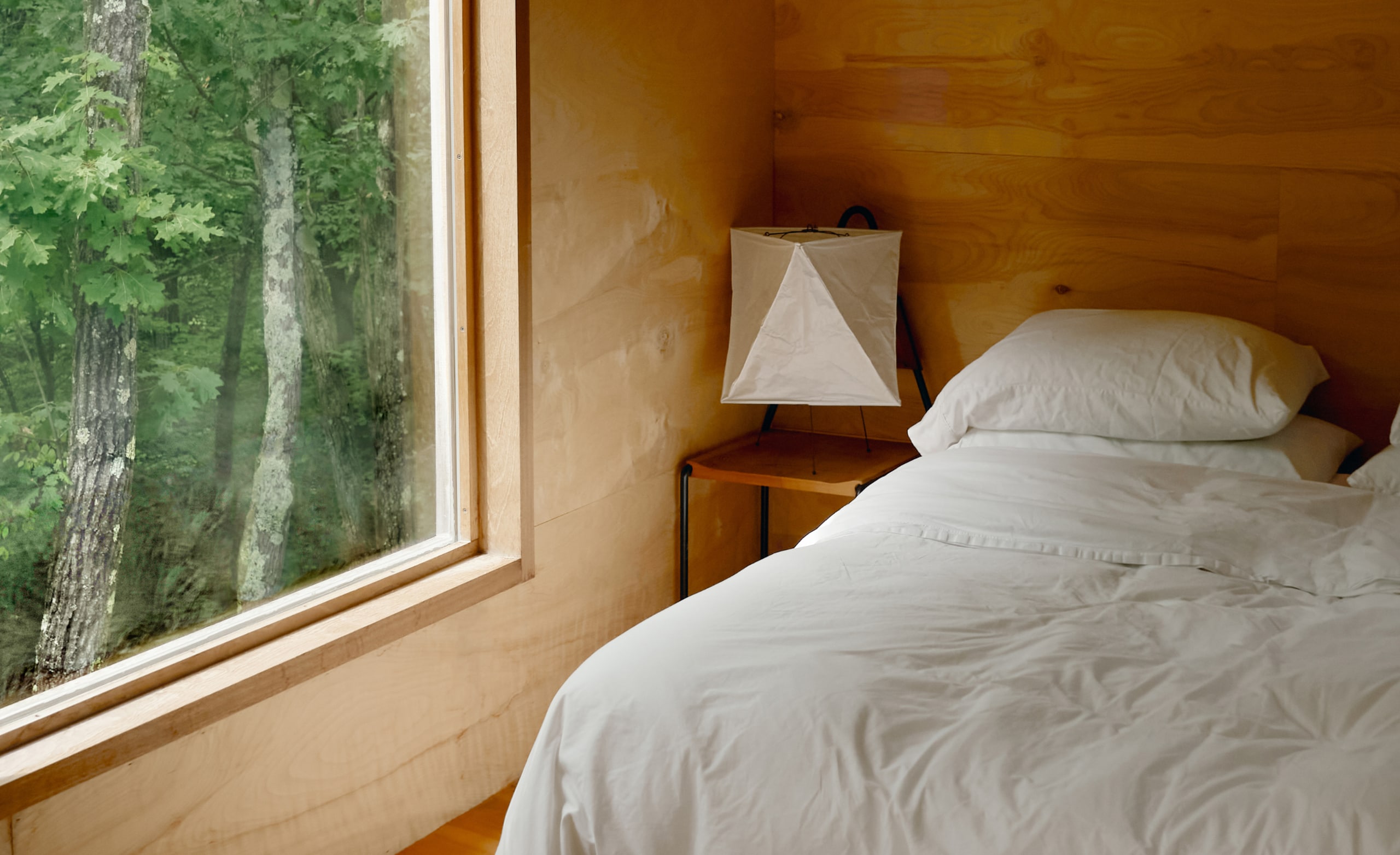 A bedroom with white sheets on a freshly made bed and a large window showing trees outside.