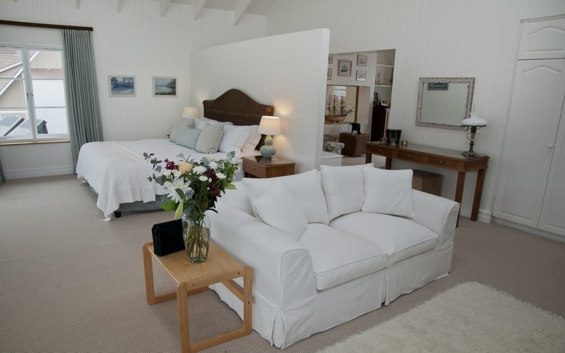 A spacious suite showcases a king-sized bed with soft linens. A white sofa is positioned beside a wooden side table holding a vase of fresh flowers. Light-colored walls brighten the room, while large windows allow natural light to illuminate the inviting space.