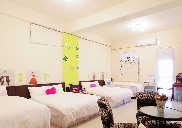 A spacious room is presented with three double beds, each adorned with white linens. Colorful wall art adds charm to the space, complemented by a small seating area featuring patterned chairs. Natural light is allowed to flow through the windows, enhancing the bright atmosphere.