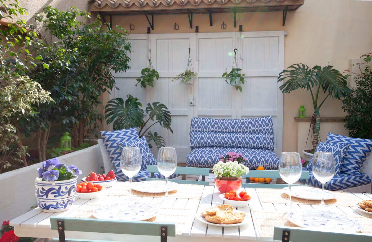 An outdoor patio area features comfortable seating against a backdrop of greenery. A table is set with glasses, fruit, and snacks, inviting al fresco dining. Decorative elements include a potted plant, enhancing the calm atmosphere of the space.