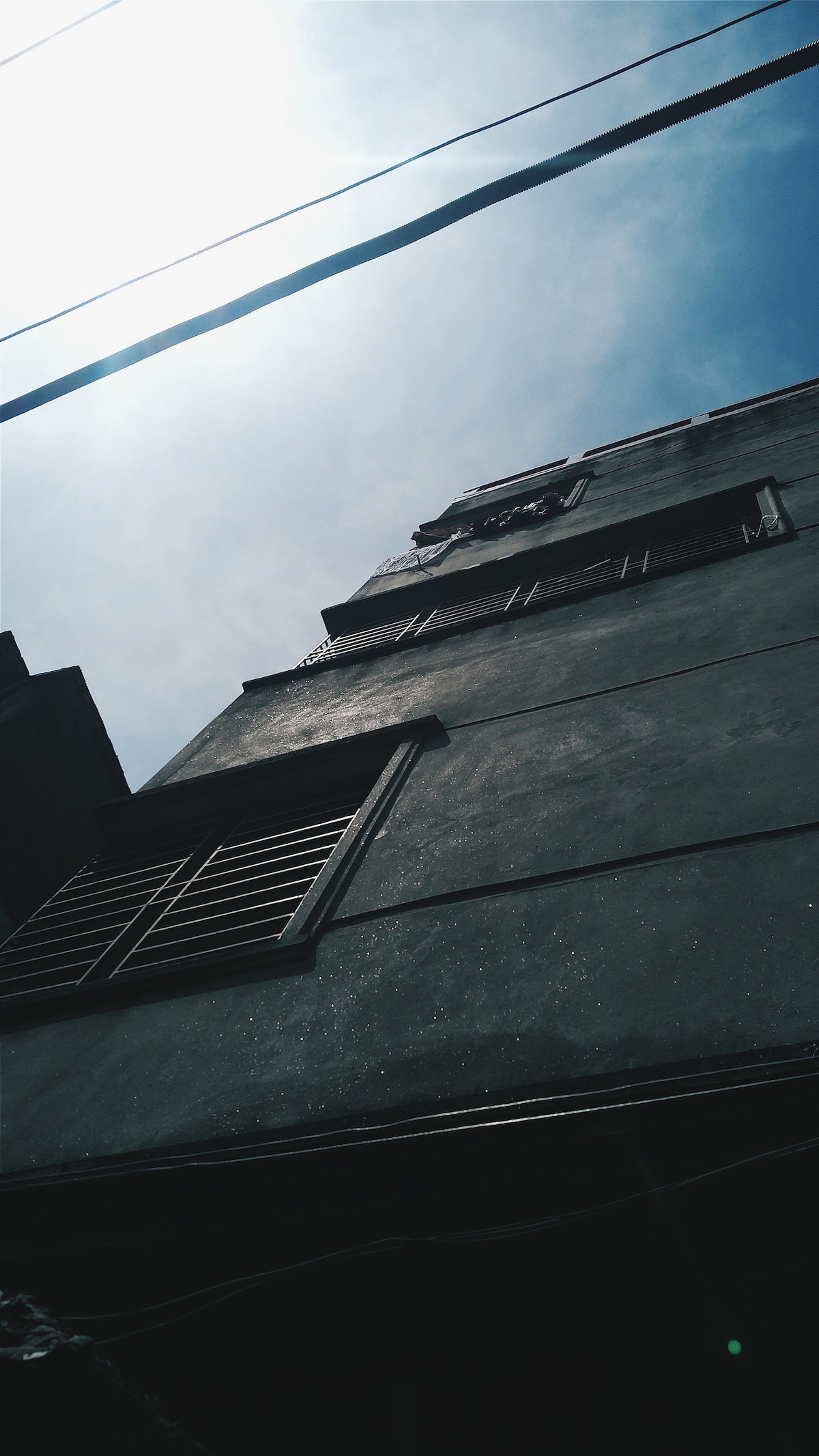 The image captures an upward view of a multi-story building, featuring several wooden shutters in a dark tone. The façade appears smooth and textured against a bright sky, with hints of sunlight reflecting off its surface.