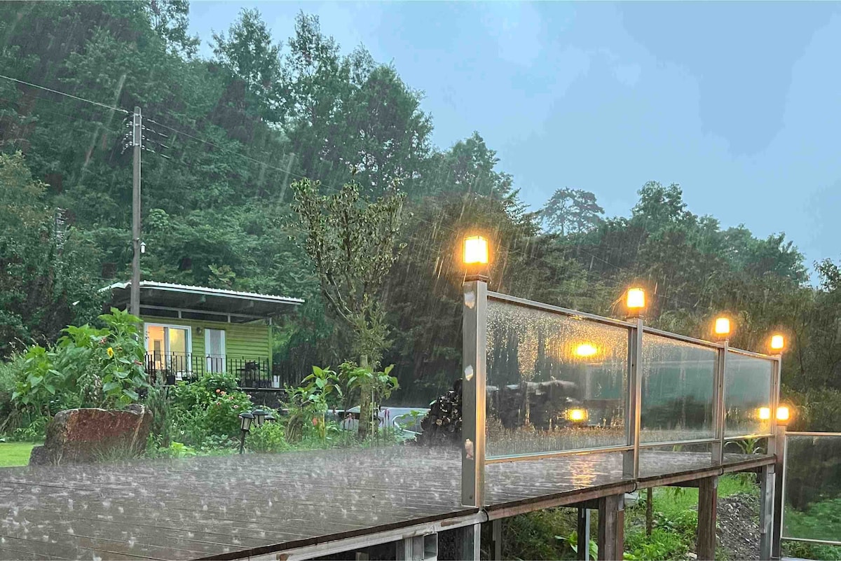A wooden pathway leads towards a charming green house surrounded by lush trees. Soft lights illuminate the path despite the rain, reflecting off the wet surface. Vegetation flanks the walkway, creating a tranquil ambiance in the natural setting.