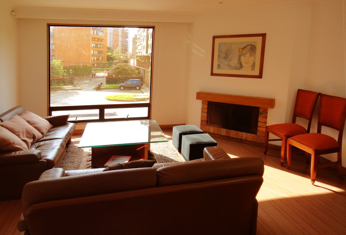 A bright living area features a large window framing a view of the outdoors. Two brown leather sofas are arranged around a glass coffee table. A stone fireplace is positioned against the wall, along with two wooden chairs placed near the wall.