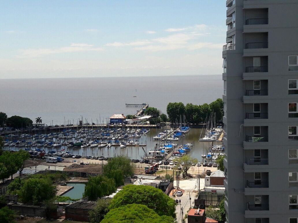 A scenic view showcases the marina lined with numerous boats, set against the calm river waters. Green trees and a small pool are visible in the foreground, while the distant pier and buildings blend into the horizon under a clear sky.