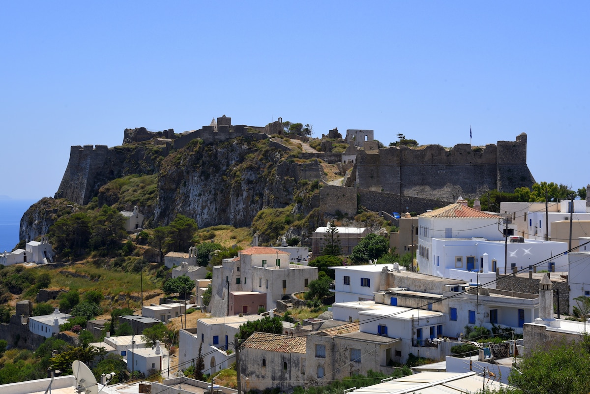 The image depicts a panoramic view of the Venetian Castle, situated on a hilltop, surrounded by the traditional white houses of Chora. The fortress walls are prominent, while the blue sky creates a vibrant backdrop, highlighting the historical significance of the site.
