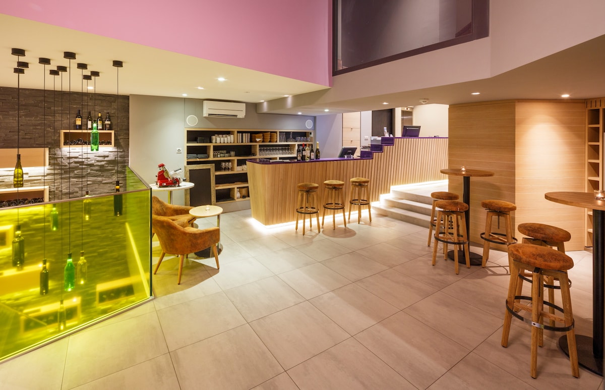 A modern bar area features a curved countertop with illuminated green accents. Stylish wood stools are arranged around high tables, with soft lighting enhancing the space. Shelves display various bottles, and an open staircase leads to a second level.
