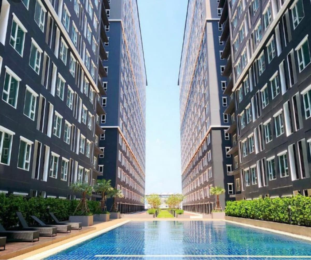 A modern building complex is framed by two adjoining structures, creating a pathway that leads to a serene, sparkling pool at the center. Lush greenery and sun loungers line the edges, enhancing the tranquil setting under a clear blue sky.
