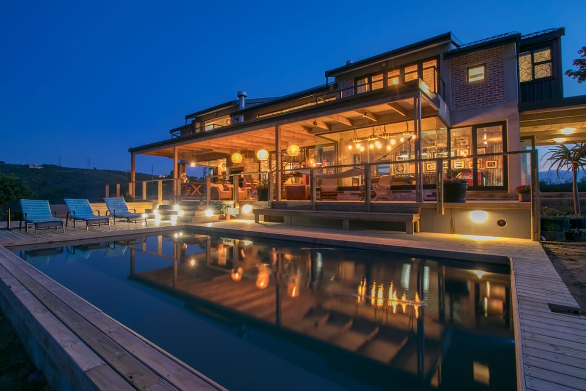 The exterior view of a two-story holiday home is shown at dusk, featuring a large wooden deck with a swimming pool reflecting the warm lights above. Comfortable seating is arranged around the deck, and the expansive windows of the home reveal inviting indoor spaces.