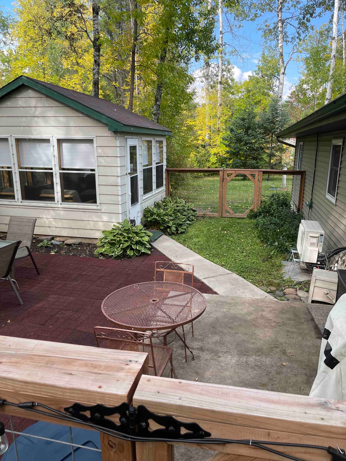 An outdoor area features a circular metal table surrounded by four chairs, set against a backdrop of lush greenery. A gazebo stands nearby, and a pathway leads through the yard, framed by trees and shrubs that provide a serene atmosphere.