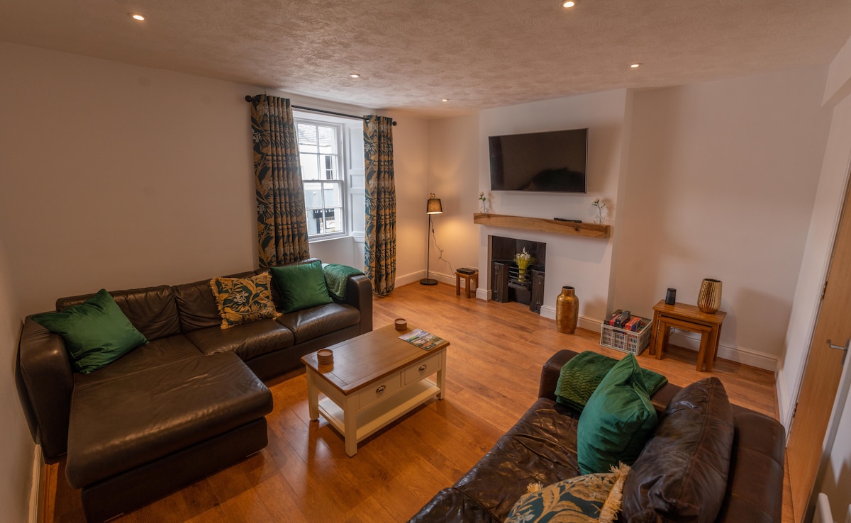 The spacious lounge features two comfortable sofas adorned with decorative cushions. A wooden coffee table sits in the center, and a flat-screen TV is mounted above a simple mantel. Natural light enters through a large window, complemented by curtains with a subtle pattern.