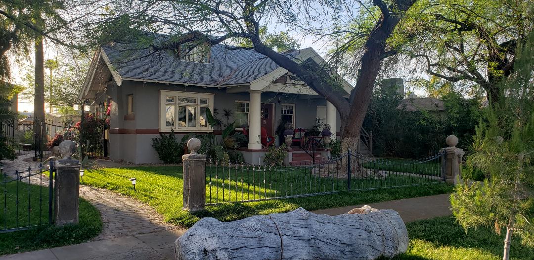 A charming historic guest house is surrounded by lush greenery and a well-maintained lawn. A pathway leads to the front porch, where potted plants add a touch of color. The house features a distinctive roof and multiple windows, enhancing its welcoming appearance.