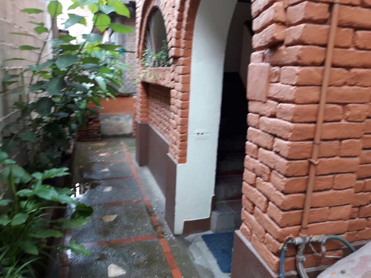 A narrow passage lined with plants leads to an entrance. The red brick walls create a rustic charm, while the pathway is tiled and slightly wet, suggesting recent rain. The archways introduce a warm atmosphere, inviting guests into the building.
