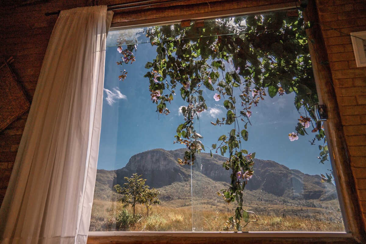 A large window reveals a scenic view of the surrounding landscape, framed by lush greenery and colorful flowers. The landscape features rolling hills under a clear blue sky, with sunlight illuminating the natural beauty of the area.
