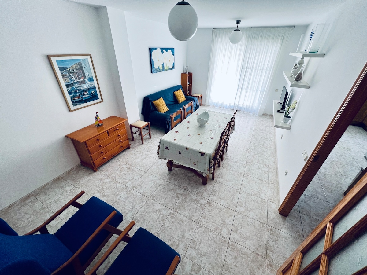 A bright dining area features a table surrounded by wooden chairs, complemented by a serene wall art piece. A green sofa with colorful cushions provides additional seating. Natural light filters through sheer curtains, and a wooden sideboard is positioned against one wall.
