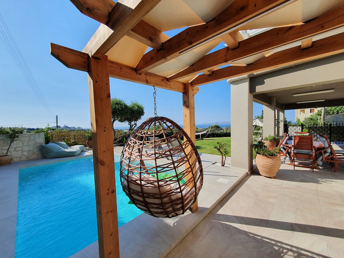 A private heated swimming pool is visible, with crystal clear water reflecting the sky. Next to the pool, a cozy hanging chair adds comfort under a wooden pergola. Green landscaping and distant views create a sense of tranquility in this outdoor space.