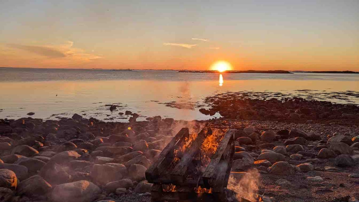A serene coastal scene showcases a tranquil sunset over the water, with the sun casting a warm glow. A fire pit sits surrounded by smooth stones, producing soft smoke that drifts into the air, enhancing the peaceful atmosphere by the beach.