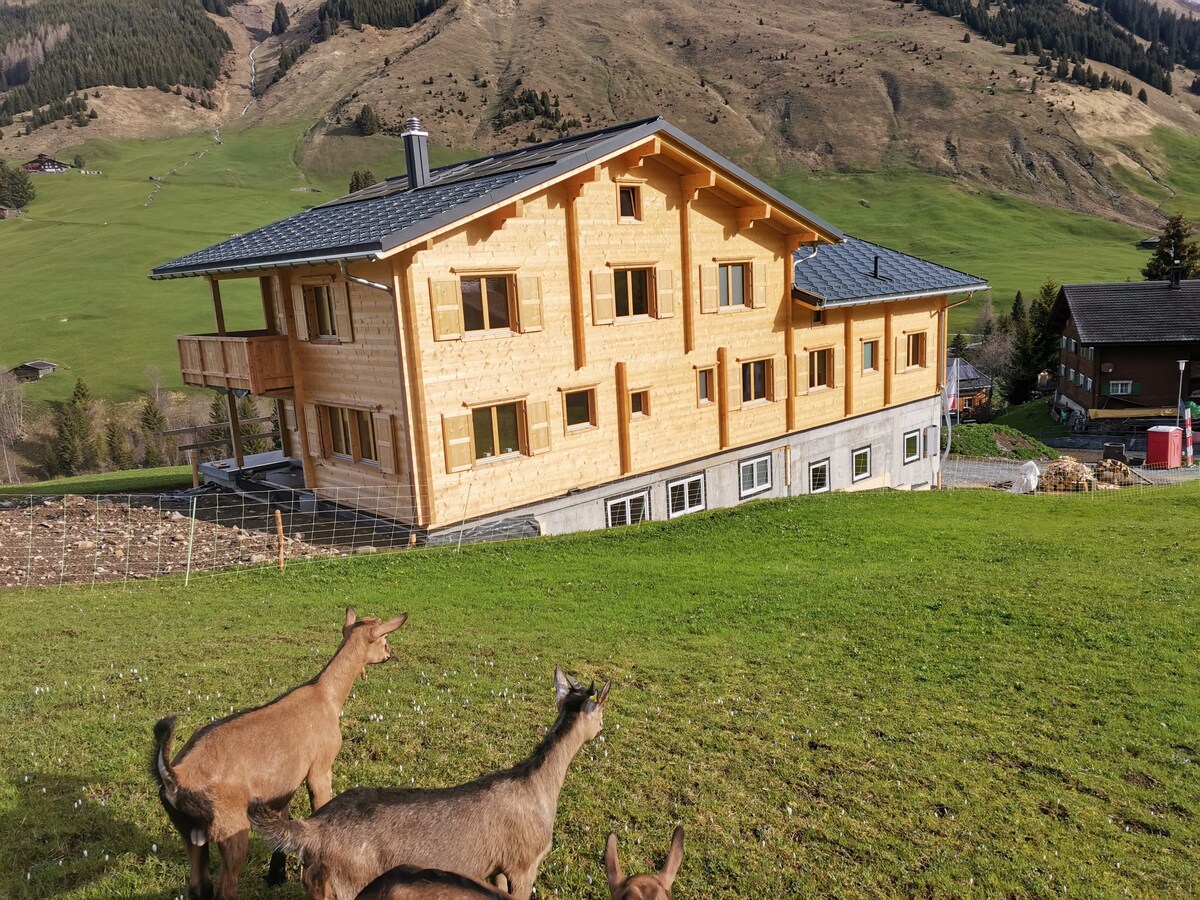 A wooden chalet-style building is situated on a grassy hillside, surrounded by mountains. The structure features multiple windows and a large balcony. In the foreground, several goats are grazing, adding a rustic charm to the tranquil landscape.