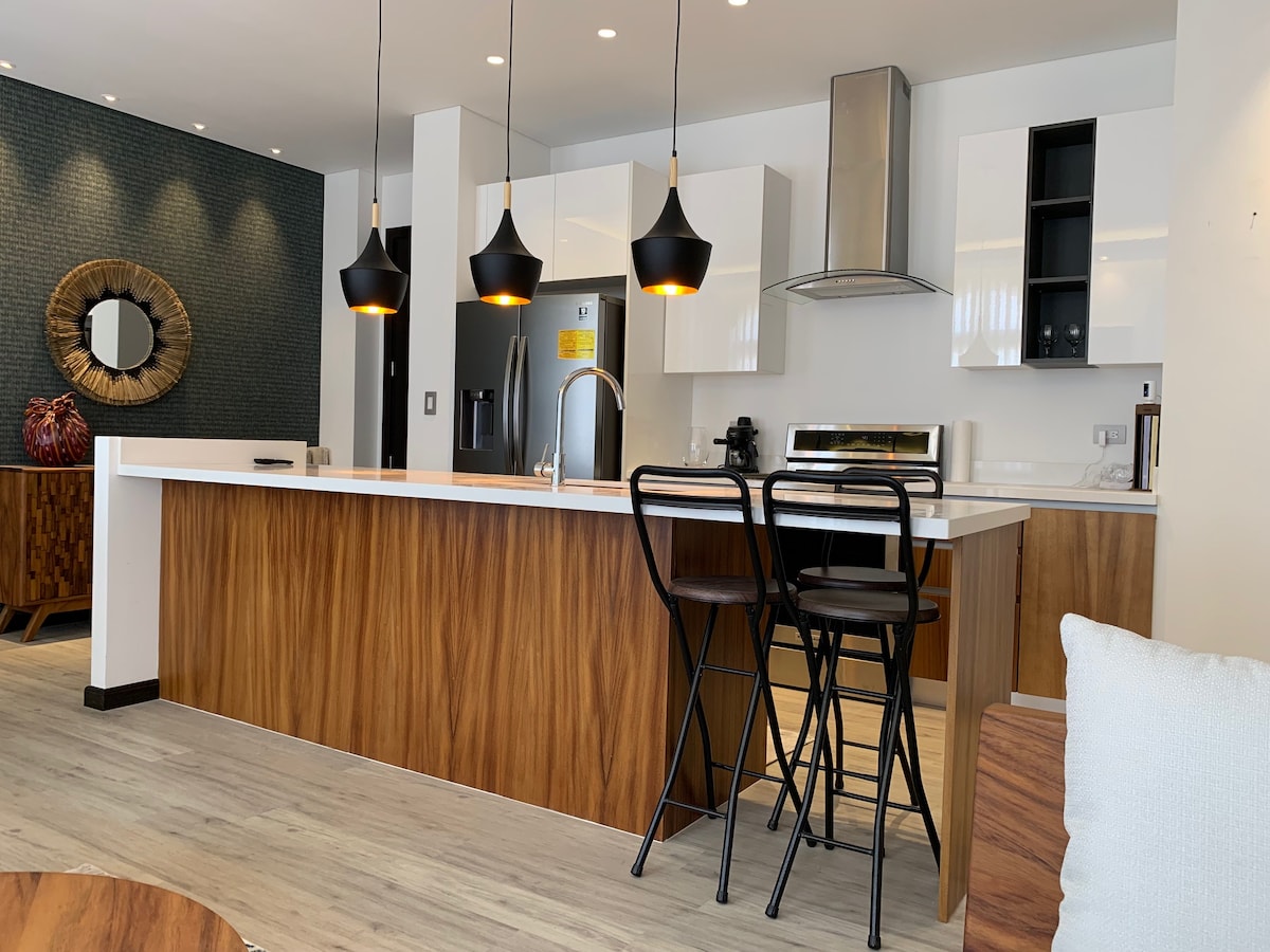 A modern kitchen is showcased with sleek white cabinets and a spacious countertop. Two high-backed black stools are positioned at the wooden breakfast bar. A stylish mirror adorns the wall, while pendant lights illuminate the space, providing a warm glow.