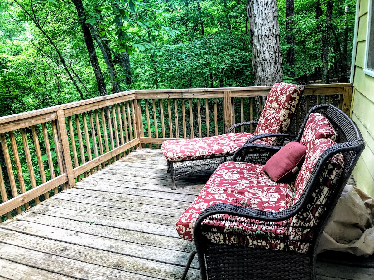 A spacious deck nestled in the trees features two comfortable lounge chairs with floral-patterned cushions, providing a serene space for relaxation. The surrounding greenery creates a peaceful atmosphere, enhancing the natural setting of the cottage.