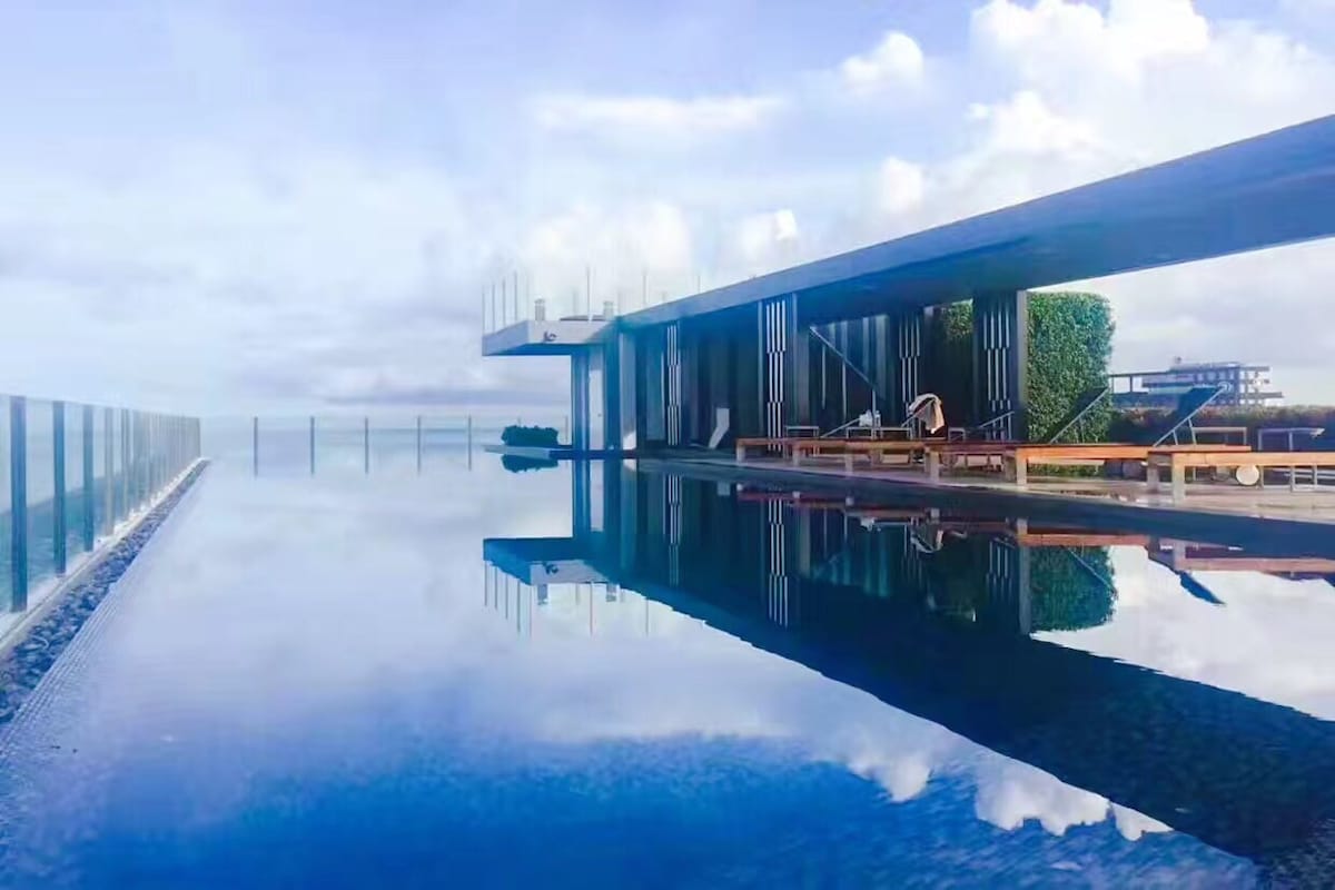 An infinity pool stretches across the image, reflecting the sky and surrounding greenery. Comfortable lounge chairs are positioned along the edge, with a modern structure visible in the background. The setting offers a panoramic view, enhancing the overall sense of tranquility.