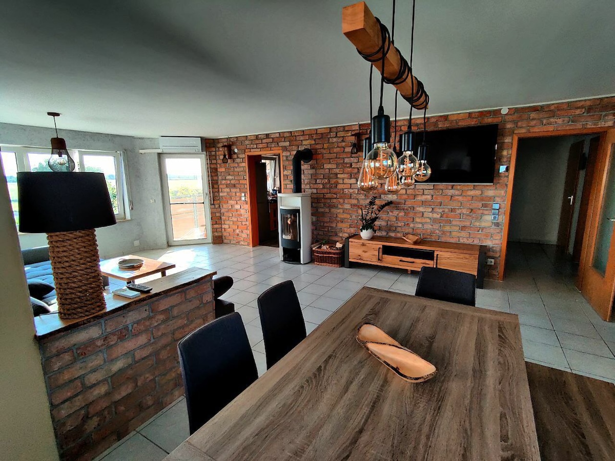 A spacious living area is highlighted by a rustic brick wall and large windows that allow natural light to fill the room. A modern dining table with black chairs sits in the center. A stylish lighting fixture hangs above, complementing the warm tones of the decor.