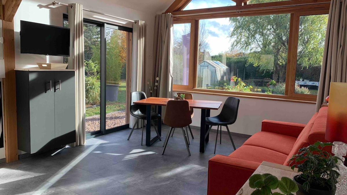 A bright living space features large glass doors that provide a view of the garden. A combination of a red sofa, a dining table for four, and modern chairs creates a functional area. Natural light is complemented by neutral-toned curtains and stylish cabinetry.
