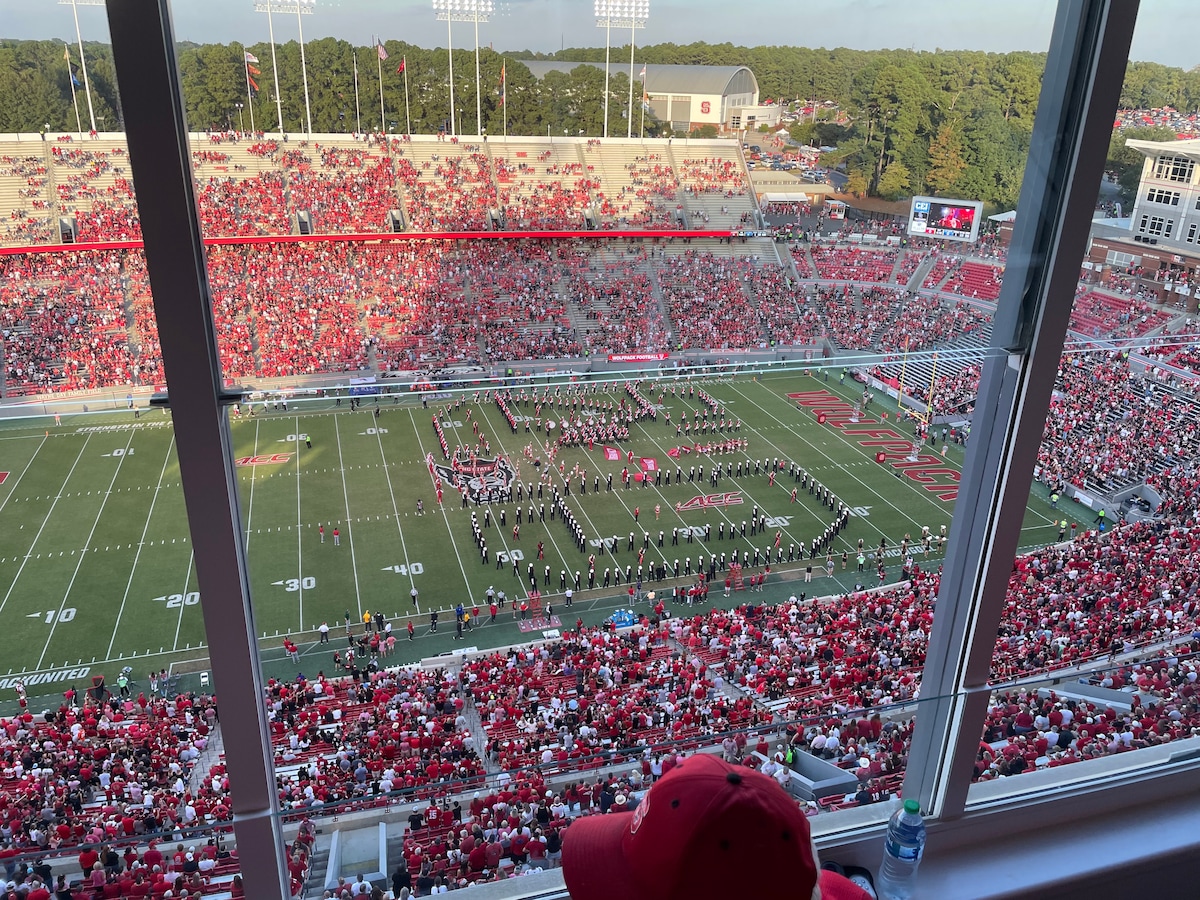 A view from a high vantage point captures a packed stadium adorned in red and white. The football field is busy with a marching band performing a formation, while spectators fill the stands, creating a lively atmosphere under the clear sky.