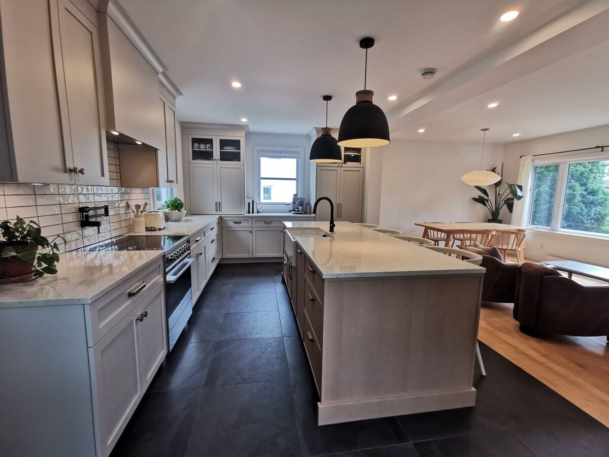 A modern kitchen features a central island with a light-colored countertop and dark cabinetry. Pendant lights hang over the island, and a large window allows natural light to illuminate the space. Adjacent seating areas are visible, creating an open and functional layout.