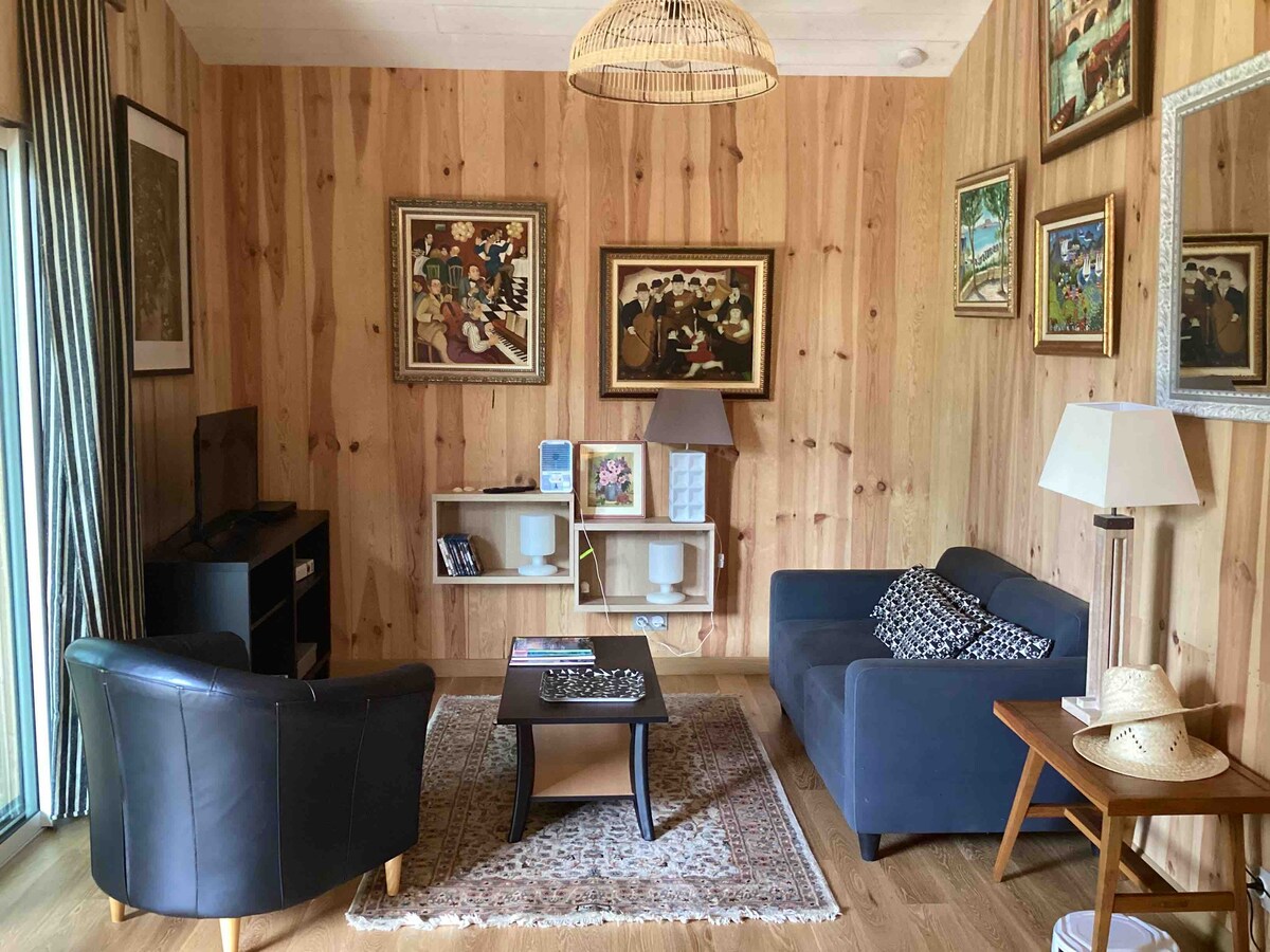A cozy living area with wooden walls and carefully arranged artwork. Seating includes a dark leather chair and a blue sofa, complemented by a coffee table. Natural light enters through large glass doors, enhancing the space's warmth. A small shelf displays various items and decor.