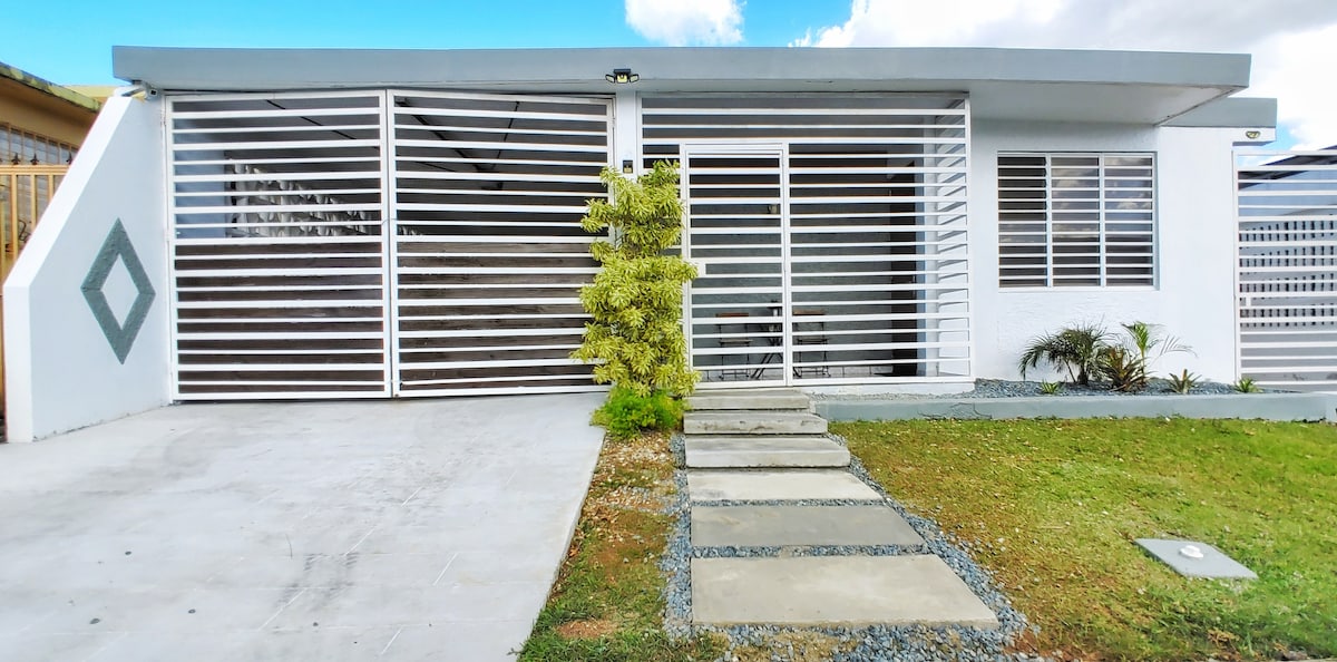 A modern exterior of a single-story house is presented, featuring horizontal white slats that provide both privacy and light. A path of stepping stones leads to the main entrance, bordered by well-maintained grass and small plants for added greenery.