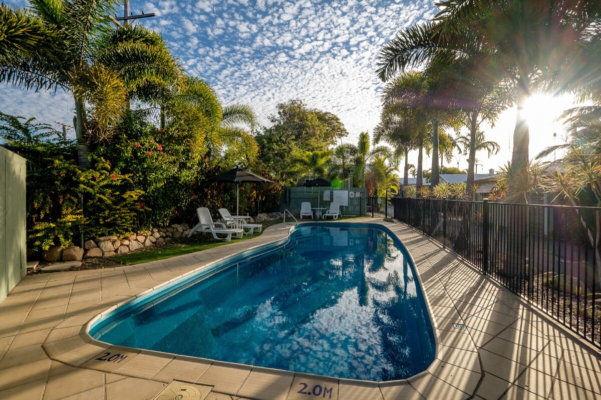 A swimming pool is bordered by tall palm trees, creating a tropical ambiance. Lounge chairs are positioned beside the pool, with sunlight reflecting off the water's surface. A gazebo is visible in the background, providing shaded seating options among lush greenery.