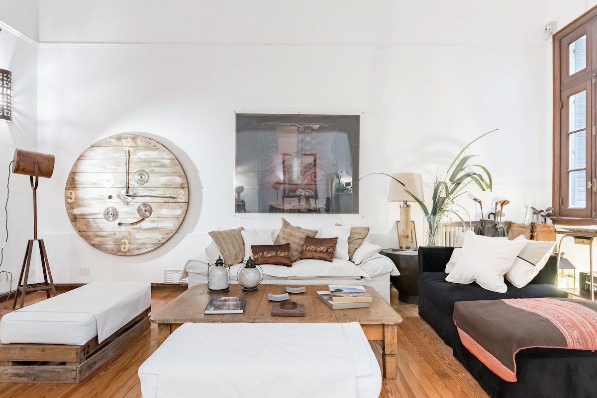 The living room features a wooden coffee table surrounded by a sofa and low sofas, providing ample seating. A large clock art piece adorns the wall, while a tall plant adds a touch of greenery. Natural light enters through the windows, enhancing the spacious feel.
