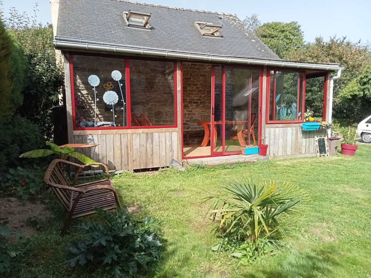 A sunlit veranda features large red-framed windows, providing views of the surrounding garden. Inside, a wooden table and chairs are positioned for dining, while planters with colorful flowers add charm to the outdoor area. A gently sloping lawn leads to a small palm plant.