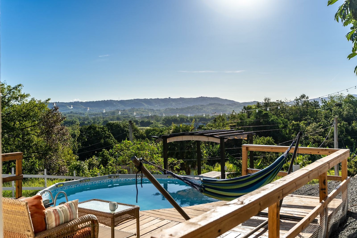 A wooden deck overlooks a tranquil pool surrounded by lush greenery. A colorful hammock hangs nearby, providing a perfect spot for relaxation. The landscape features rolling hills under a clear blue sky, inviting a sense of calm and connection with nature.