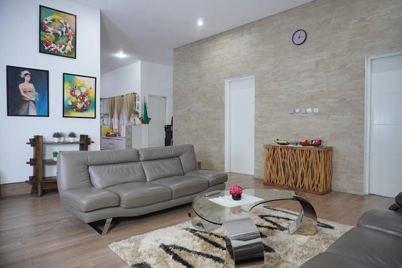 A spacious living area features a contemporary grey leather sofa, arranged around a sleek glass coffee table. A patterned rug lies beneath, complemented by artwork on the walls. A clock is mounted above, indicating the time, while doors lead to other parts of the space.