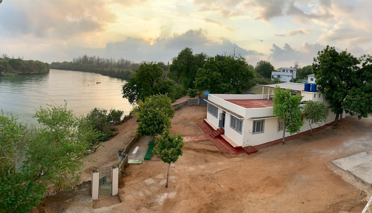 A serene view of the Takaungu Creekside villa is captured, with soft morning light illuminating the exterior. Lush green trees frame the property, while the tranquil water reflects the sky. A spacious terrace leads to the creek, enhancing the peaceful atmosphere.