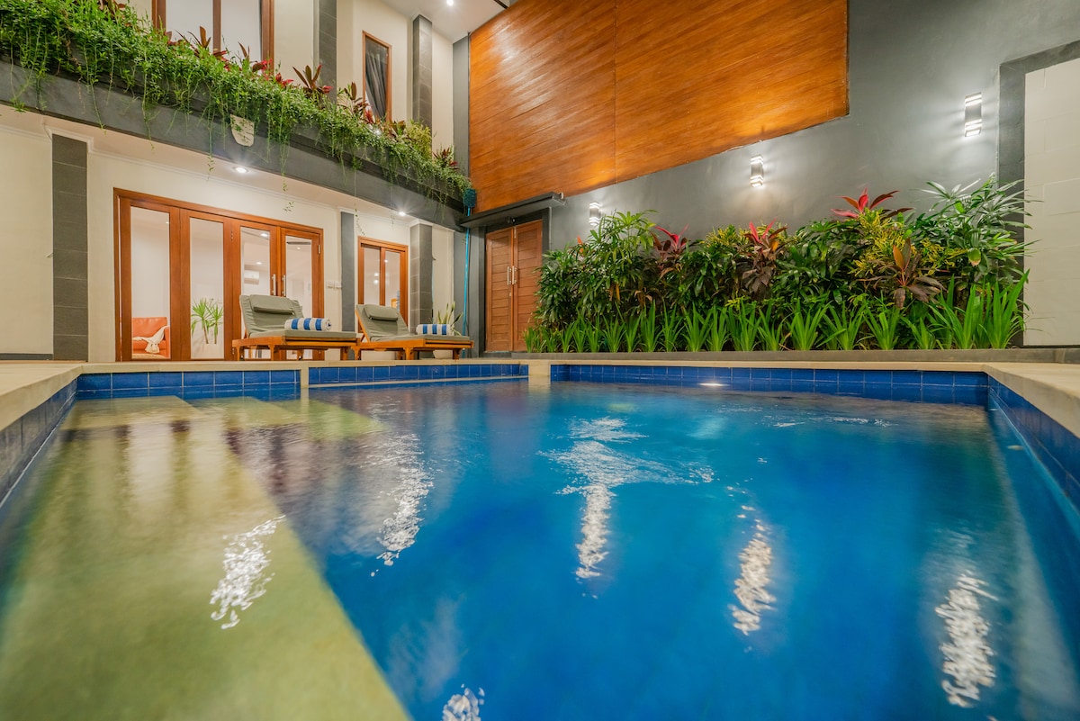 A sparkling blue pool occupies the center of the image, with a smooth and tiled surrounding. Lush tropical greenery frames the area, adding a vibrant touch. Two comfortable lounge chairs are positioned nearby, under warm lighting that highlights the modern architectural features of the villa.
