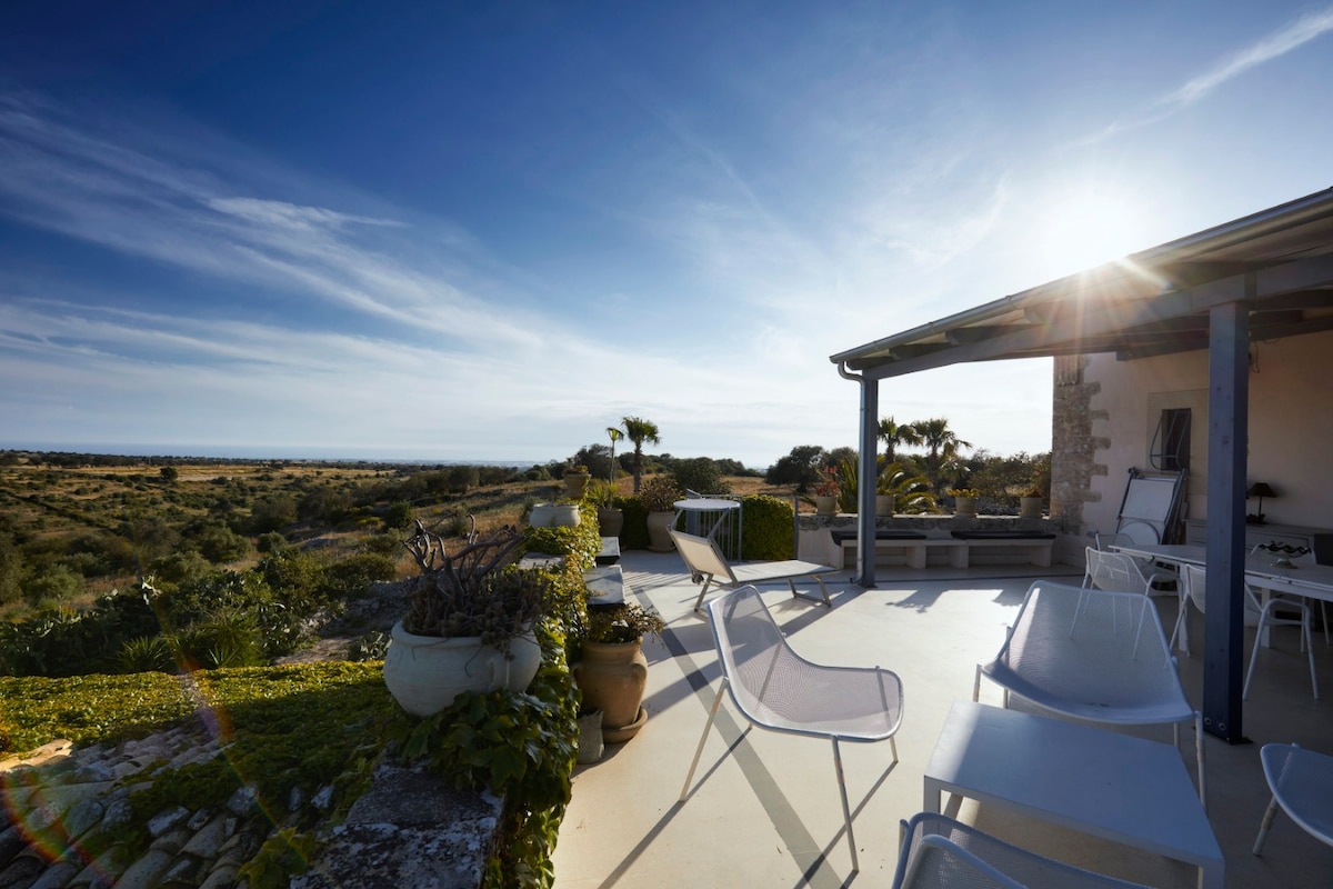 A sunlit terrace is shown with several white lounge chairs, offering views of the expansive landscape. Mediterranean plants are visible in pots, with the distant horizon revealing a glimpse of the sea under a clear blue sky.