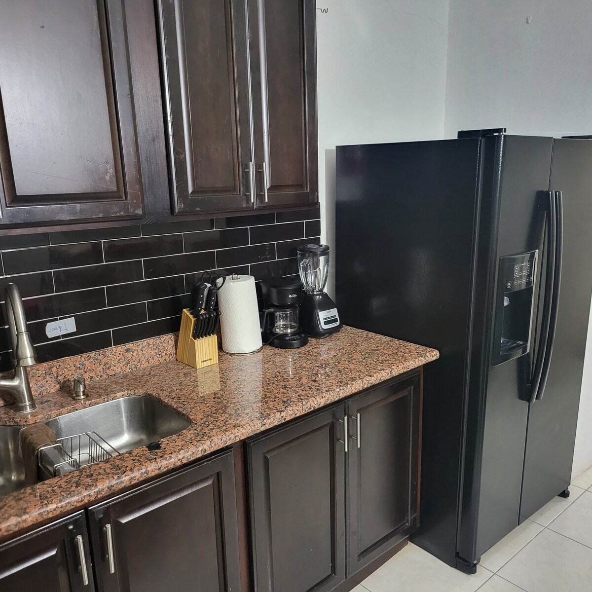 The kitchen features dark wood cabinetry complemented by a granite countertop. A stainless steel refrigerator stands alongside a blender and coffee maker. A stainless steel sink with a modern faucet is situated against a backdrop of black tiled walls.