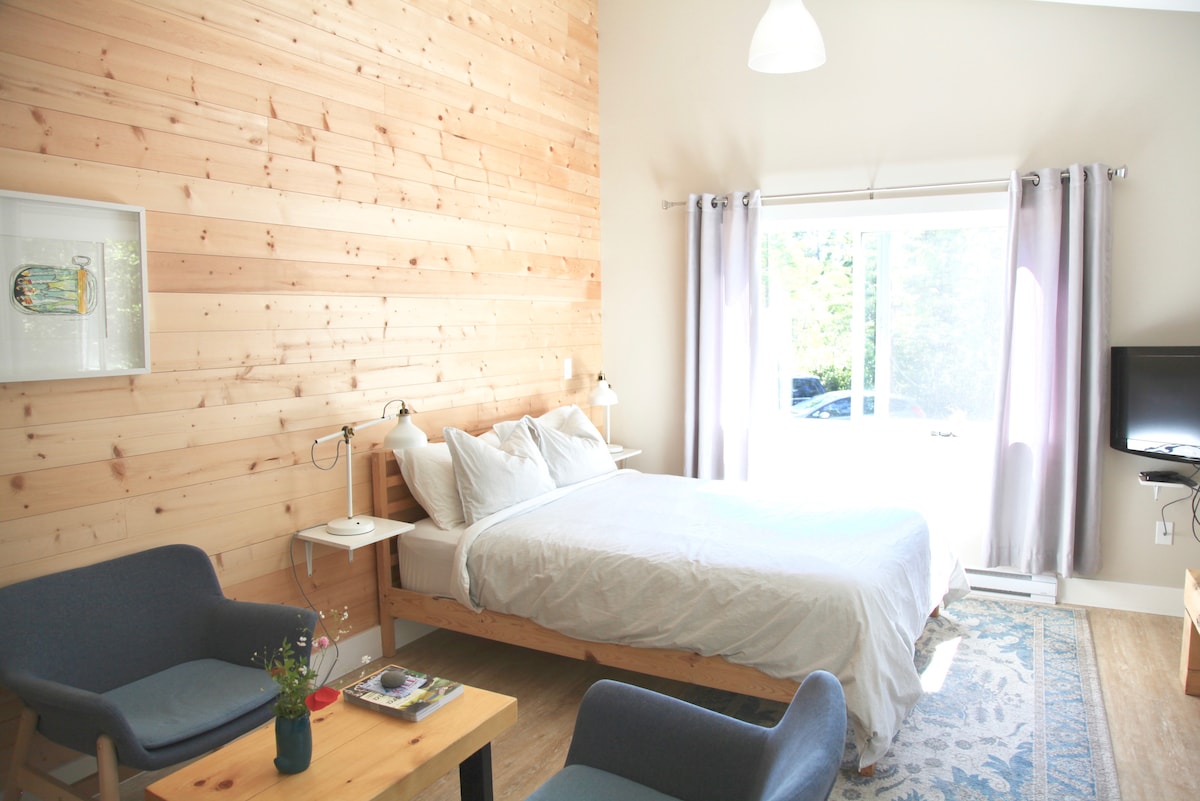 A light and airy bedroom features a spacious bed dressed in white linens, flanked by bedside lamps. Two blue chairs are positioned near a wooden table, and a mounted television is visible on the wall. Large windows allow natural light to fill the room.