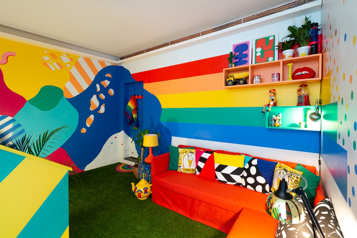 A vibrant living space is filled with colorful walls featuring bold patterns and stripes. A red sofa is complemented by eclectic cushions, while a small lamp and shelves display playful decor. The floor is covered in green carpet, enhancing the cheerful atmosphere.