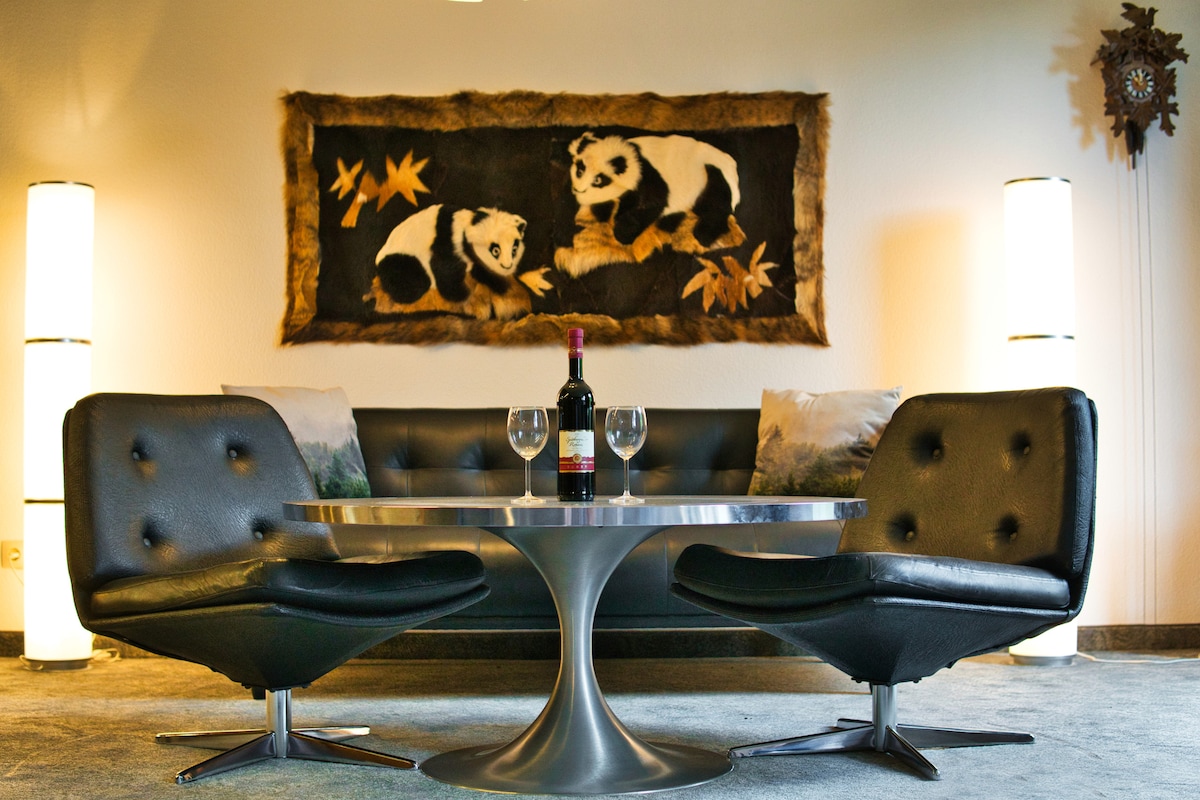 A sleek black sofa is paired with two modern chairs around a round metallic table, set with two wine glasses and a bottle of wine. A large wall hanging of panda bears adds a touch of character to the space, illuminated by floor lamps.