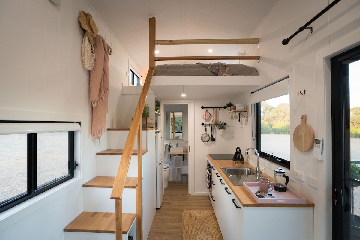 An open-plan interior is highlighted by a wooden staircase leading to a lofted sleeping area. A minimalist kitchenette features modern appliances and simple decor. Natural light enters through large windows, illuminating the cozy living space adorned with functional elements and a warm-toned area rug.