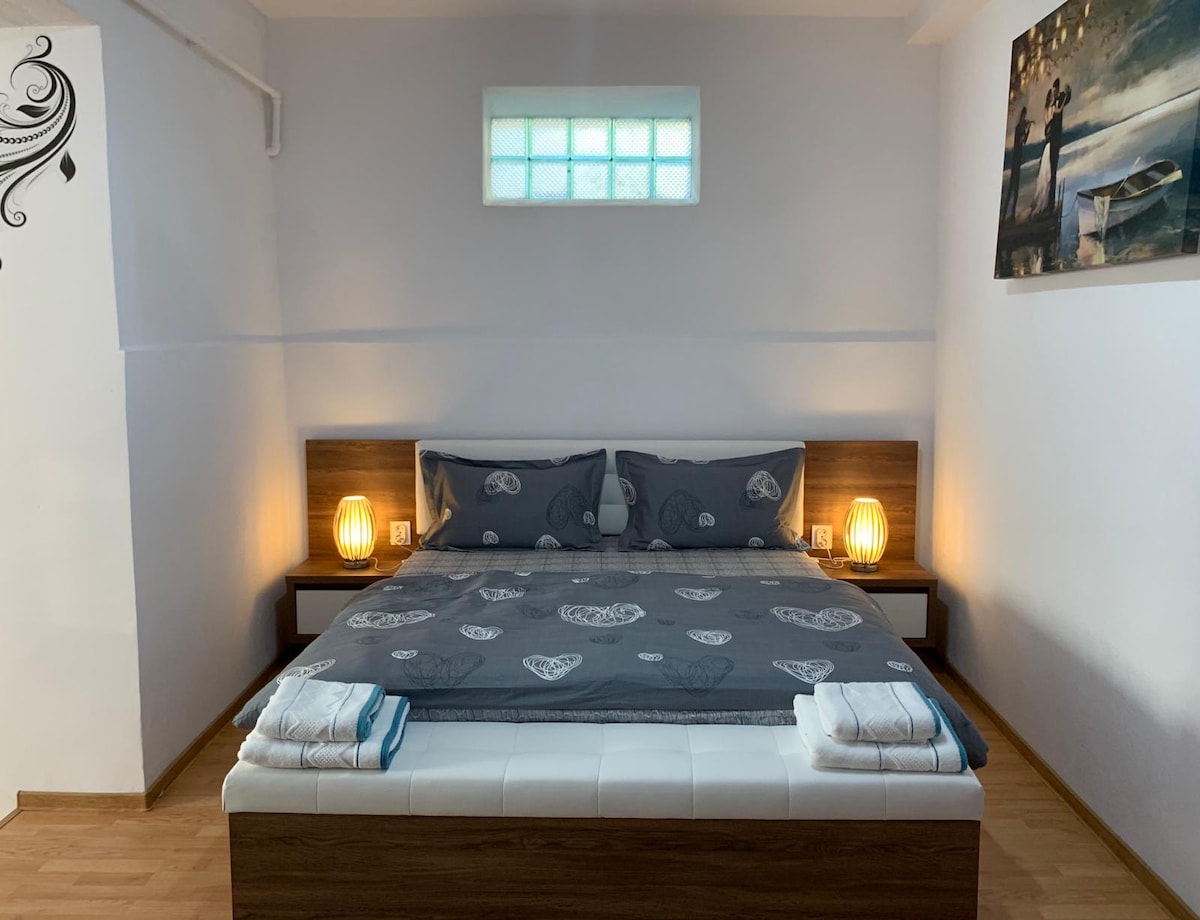 A spacious sleeping area is outfitted with a large bed featuring dark bedding and decorative throw pillows. Contemporary bedside lamps cast a soft glow, and the light from a small window adds warmth. Neatly folded towels are arranged at the foot of the bed.